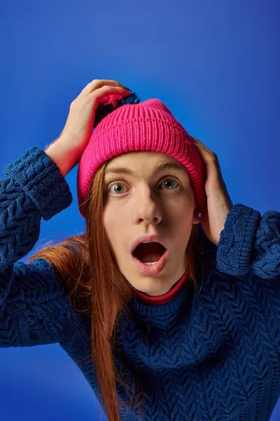 A young man with long red hair reacts in shock while wearing a bright pink beanie and blue sweater — Stock Photo