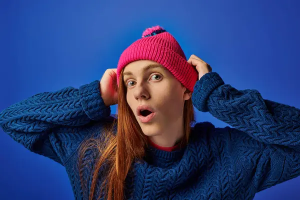 A young man with long red hair, surprised, wears a pink beanie against a blue backdrop. — Stock Photo