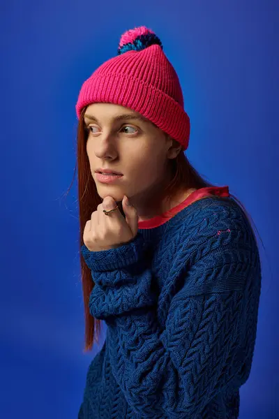 Un joven con el pelo rojo largo en un gorro brillante muestra emociones profundas contra un telón de fondo azul. - foto de stock