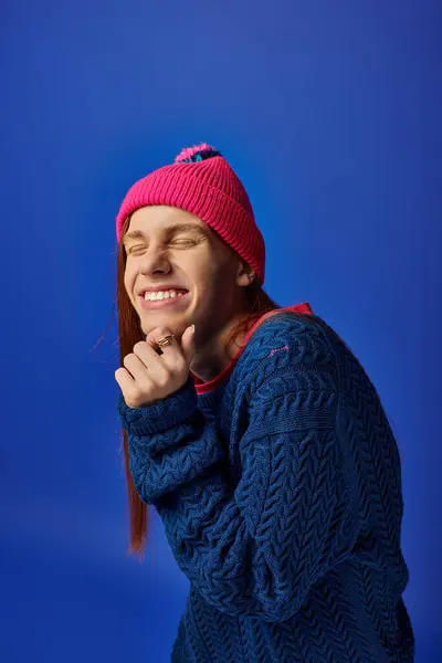 A young man with long red hair and a pink beanie shows a joyful emotion against a blue background. — Stock Photo