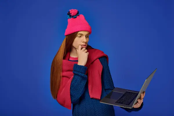 Young man with long red hair reflects while holding a laptop in a vibrant blue setting. — Stock Photo
