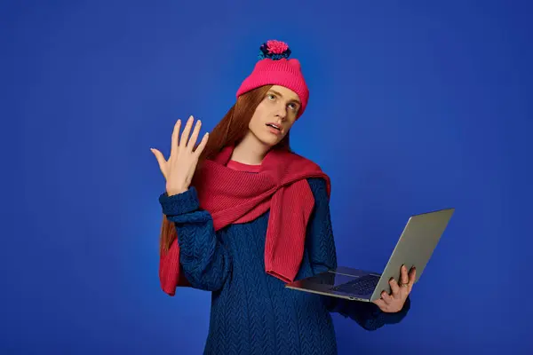 Joven emocional con el pelo largo y rojo en un gorro brillante y suéter se involucra con un ordenador portátil. - foto de stock
