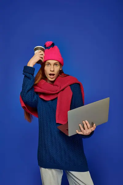 Emotional young man with long red hair holds a laptop and coffee cup, displaying shock. — Stock Photo