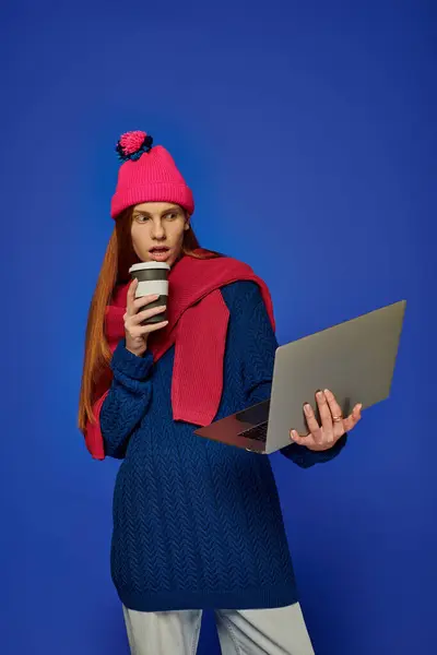 A young man with long red hair holds coffee and uses his laptop, expressing deep emotions. — Stock Photo