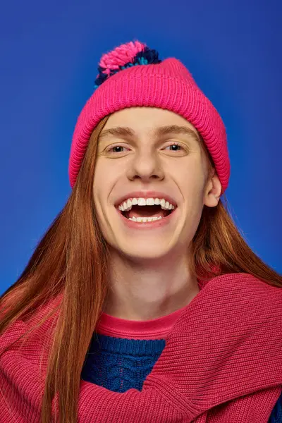 Emotional young man with long red hair laughs while wearing a vibrant pink hat and sweater. — Stock Photo