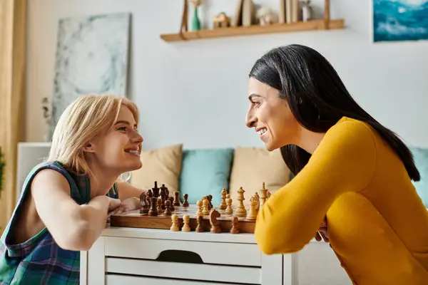 Две молодые женщины улыбаются и смеются, играя в шахматы в своем стильном жилом пространстве — стоковое фото