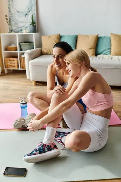 Two young women share a moment of laughter while sitting on yoga mats at home. — Stock Photo