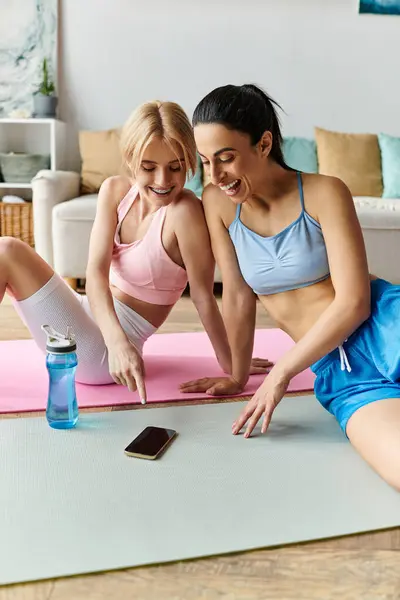 Two young women bond while checking their workout progress on a smartphone at home. — Stock Photo