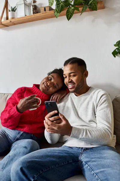 A happy couple cuddles in warm sweaters, enjoying quality time together at home on their phone. — Stock Photo