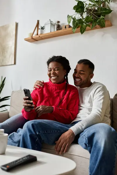 Joyful couple cuddles on the couch, sharing laughter while checking their phone in winter. — Stock Photo
