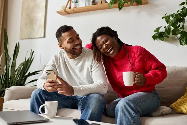 Un couple afro-américain joyeux partage le rire tout en se relaxant sur leur canapé, enveloppé dans la chaleur. — Photo de stock