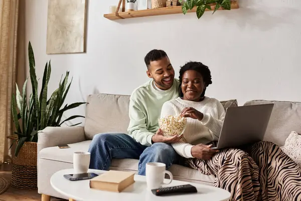 An African American couple laughs on the couch, sharing popcorn and working on a laptop. — Stock Photo