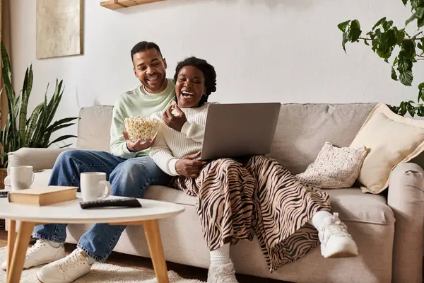 Cozy couple laughs together while sharing popcorn and watching a movie in their living room. — Stock Photo