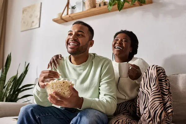 Una coppia felice si rilassa sul divano in maglioni accoglienti, condividendo risate e spuntini insieme. — Foto stock