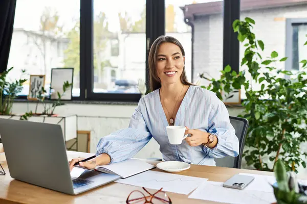 Beautiful brunette smiles while sipping coffee, engaged in work at her desk surrounded by plants. — Stock Photo