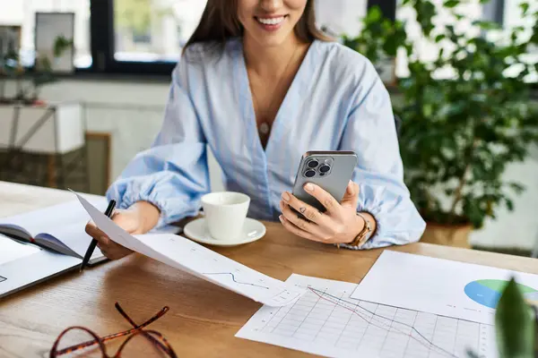 Brunette woman smiles as she checks her phone, surrounded by papers and a warm cup of coffee. — Stock Photo