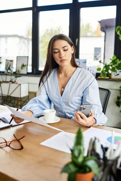Brunette woman seated at a desk, checking her smartphone while enjoying a cup of coffee. — Stock Photo