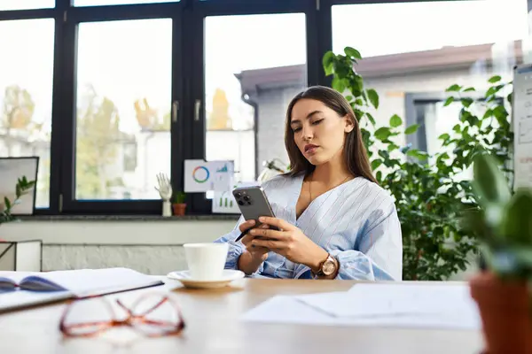 Junge Frau mit brünetten Haaren checkt ihr Smartphone, während sie ein warmes Kaffeegetränk genießt. — Stockfoto