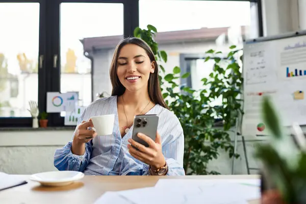 Улыбающаяся женщина с чашкой кофе вступает в контакт со своим смартфоном в светлом офисе — стоковое фото