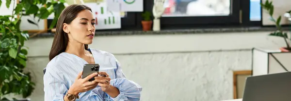 A beautiful brunette woman sits in a stylish office, focused on her smartphone amidst lush plants. — Stock Photo