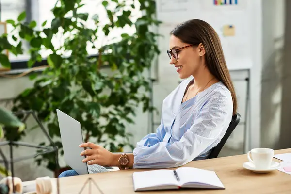 Brunette woman wearing glasses smiles while using her laptop in a lively workspace — Stock Photo