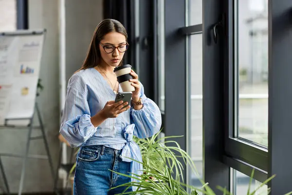 Beautiful brunette woman stands by the window, sipping coffee while texting on her phone. — Stock Photo
