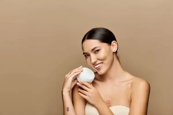Jeune femme brune met en valeur sa peau rayonnante tout en tenant un récipient de soins de la peau. — Photo de stock