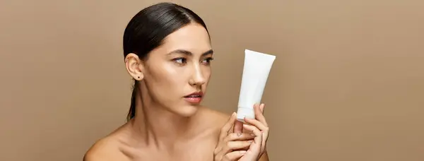 Beautiful brunette woman examines a skincare product in a softly lit, neutral toned environment. — Stock Photo