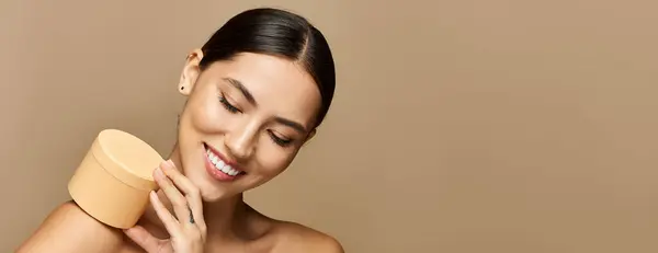 Beautiful brunette woman smiles while holding a skincare product against a neutral backdrop — Stock Photo