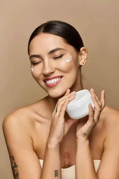 A young beautiful woman smiles while applying cream to her face, enjoying a skincare ritual. — Stock Photo