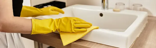 Bright kitchen environment where a beautiful young woman cleans a sink with yellow gloves. — Stock Photo
