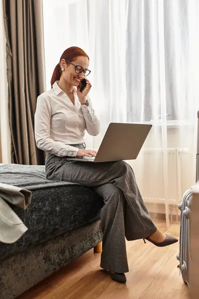 Una giovane donna si siede sul letto, con fiducia su una chiamata e lavorare sul suo computer portatile. — Foto stock