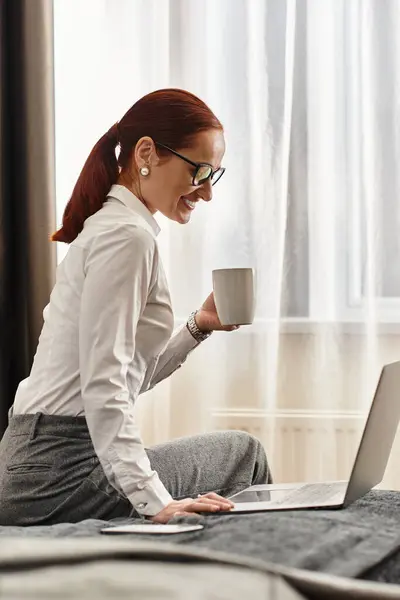 Jovem sorri enquanto trabalhava em seu laptop, segurando uma xícara de café em um quarto acolhedor. — Fotografia de Stock