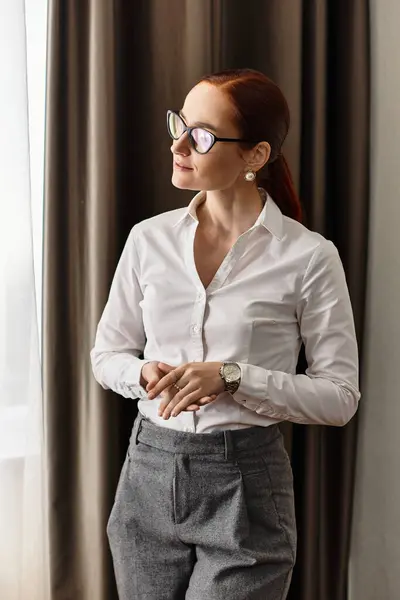 Beautiful young woman stands by the window, wearing a sophisticated outfit and lost in thought. — Stock Photo
