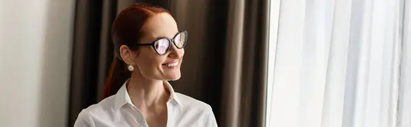 Young woman with glasses smiles brightly as sunlight filters through her window curtains — Stock Photo