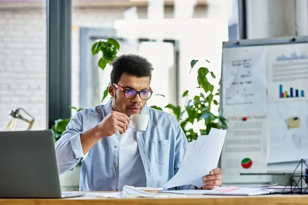 Individuo enfocado bebiendo café mientras revisa documentos en un ambiente de oficina alegre. - foto de stock