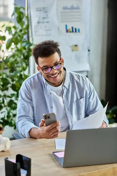 A focused man smiles at his phone while sitting at a desk surrounded by greenery. — Stock Photo