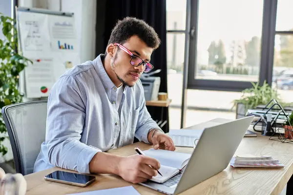 A focused man writes notes while using a laptop in a well lit office space. — Stock Photo