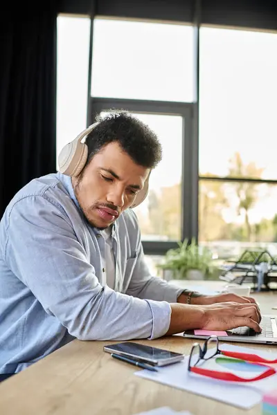 Bello uomo si concentra sul suo computer portatile, circondato da note e dispositivi mentre indossa le cuffie. — Foto stock