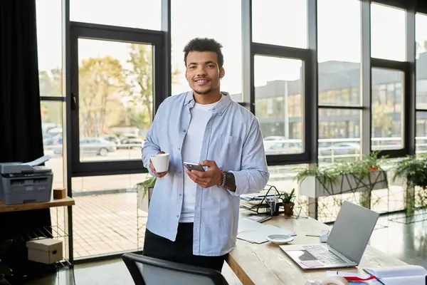 A stylish man stands in a bright office space, sipping coffee and using his smartphone. — Stock Photo