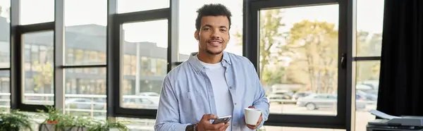 Handsome man holds a cup and smartphone, smiling in a lively, sunlit cafe. — Stock Photo