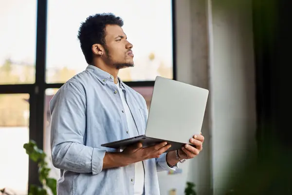 A handsome man holds a laptop while deep in thought in a sunlit space with plants. — Stock Photo
