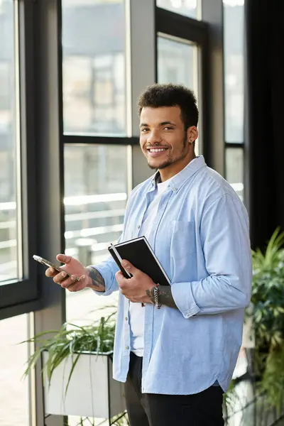 A confident man stands by large windows, smiling while holding a phone and notebook. — Stock Photo