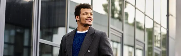 Encantador hombre vestido con abrigo elegante pasea por edificios de vidrio modernos bajo cielos azules claros. - foto de stock