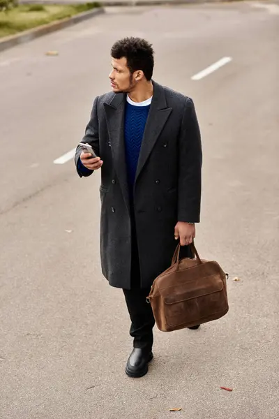 Un hombre de moda pasea por un camino urbano, revisando su teléfono mientras lleva una bolsa de cuero. — Stock Photo