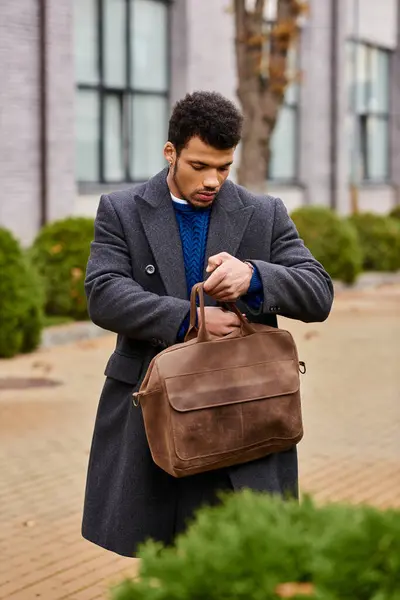 A fashionable young man retrieves items from his brown bag while walking through a city street. — Stock Photo