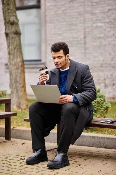 A stylish young man sips coffee and focuses on his laptop in a serene outdoor setting. — Stock Photo
