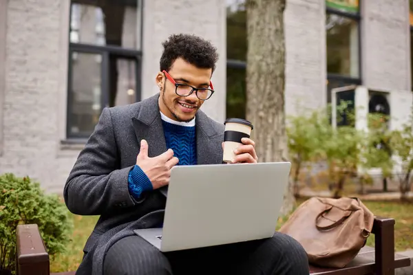A handsome man casually smiles while sipping coffee and engaging with his laptop outdoors. — Stock Photo