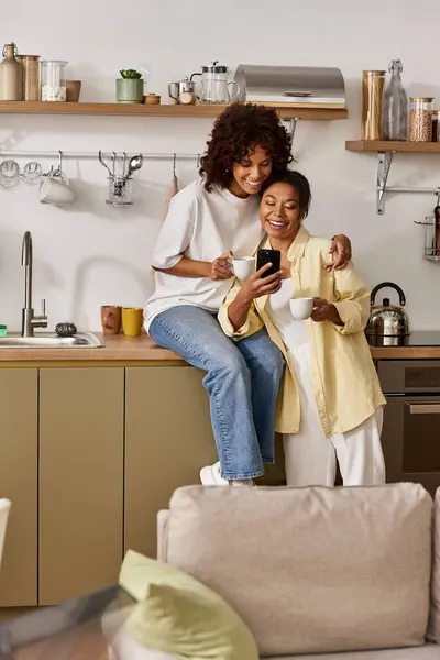 Two young women share a joyful moment while looking at a phone in their stylish kitchen. — Stock Photo