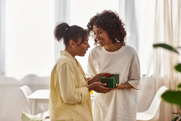 Two young women share a heartfelt moment with a gift in a bright, modern apartment. — Stock Photo